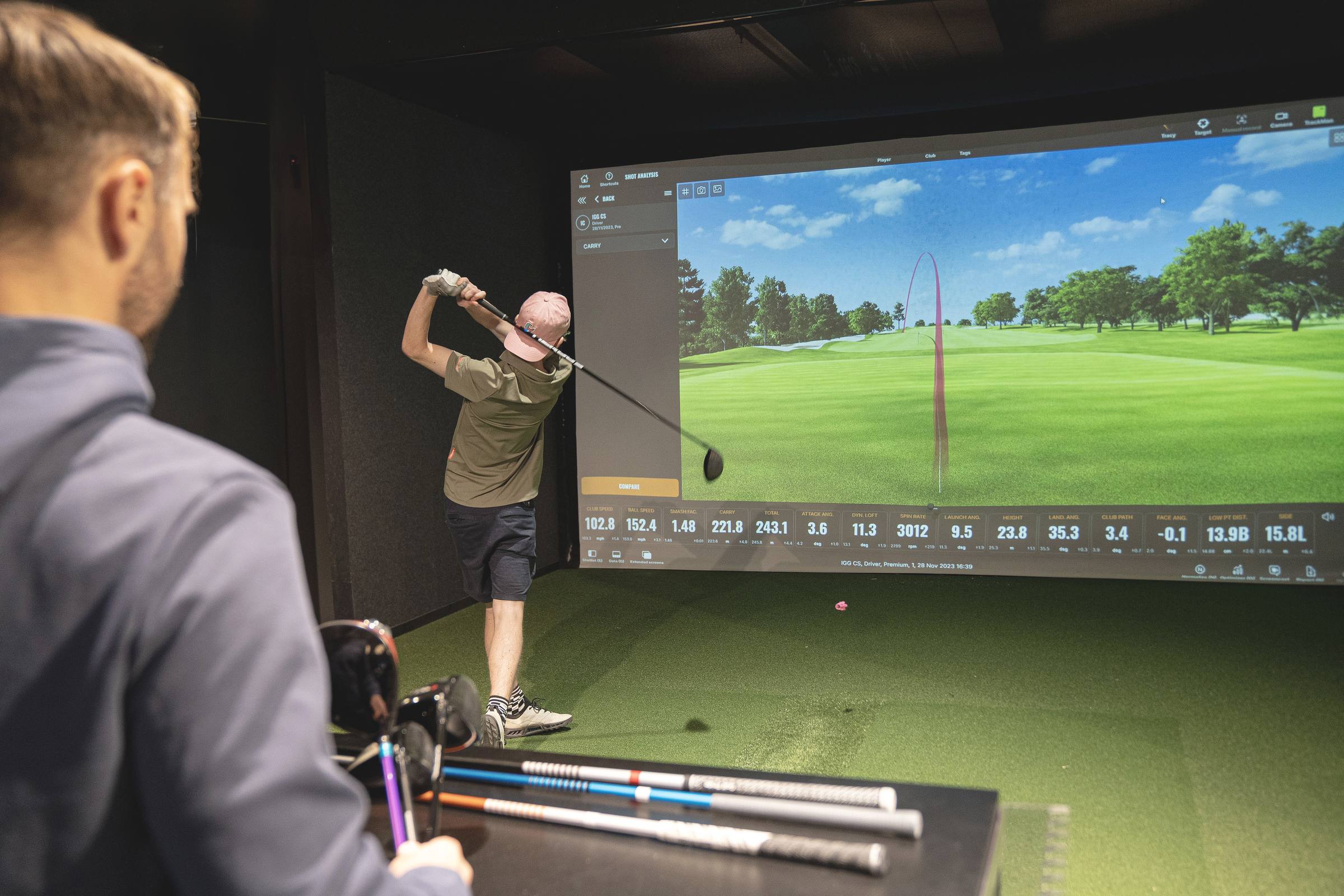 En golfare provar en driver i simulatorn medan en annan person följer slaget.